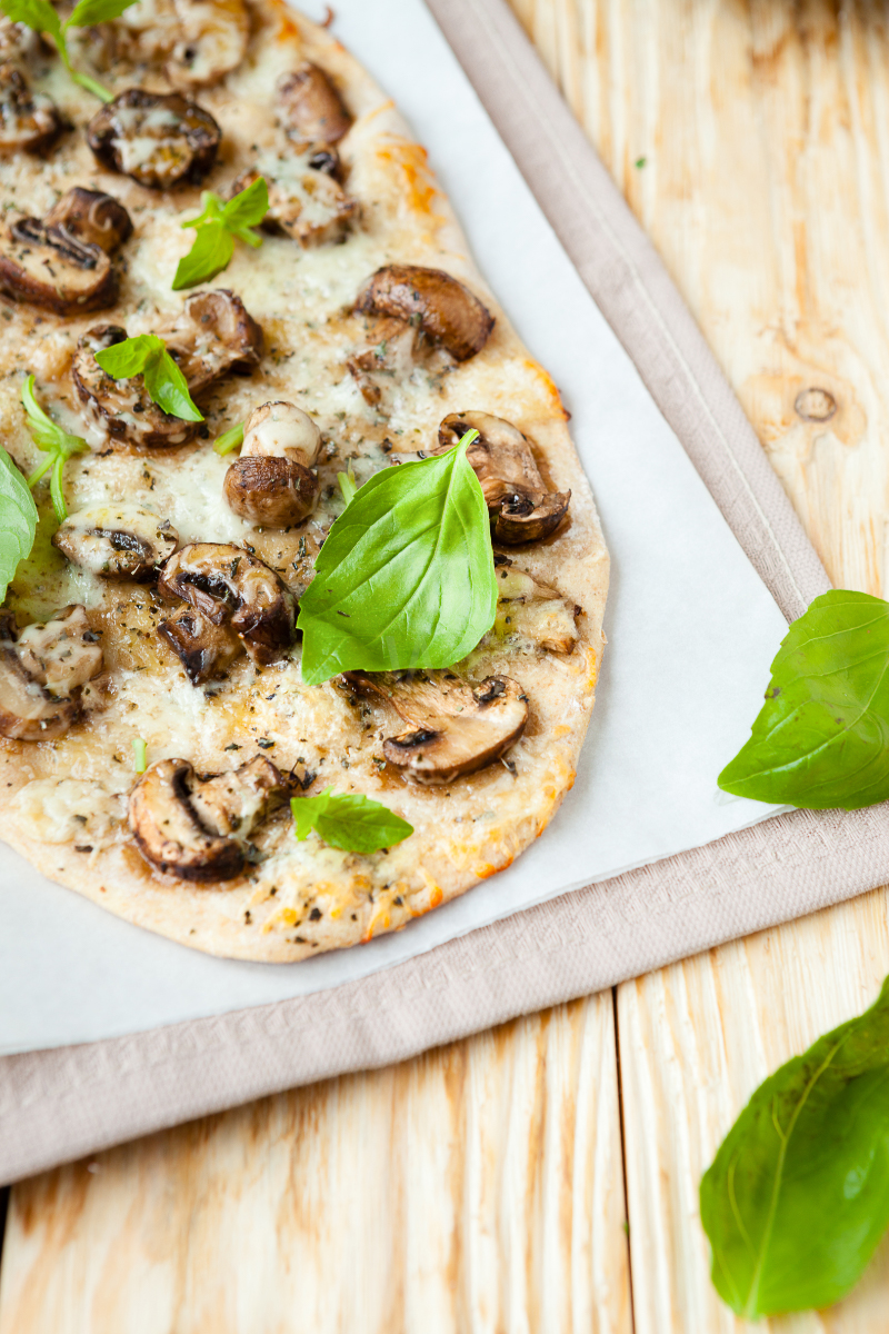 Close-up of crispy mushroom mozzarella pita flatbread with melted cheese, browned mushrooms, dried herbs, and fresh basil leaves on a light wooden table.
