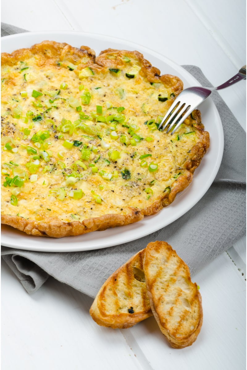 Close-up of zucchini mozzarella omelette topped with scallions and cracked black pepper, served on a white plate with grilled bread on the side.