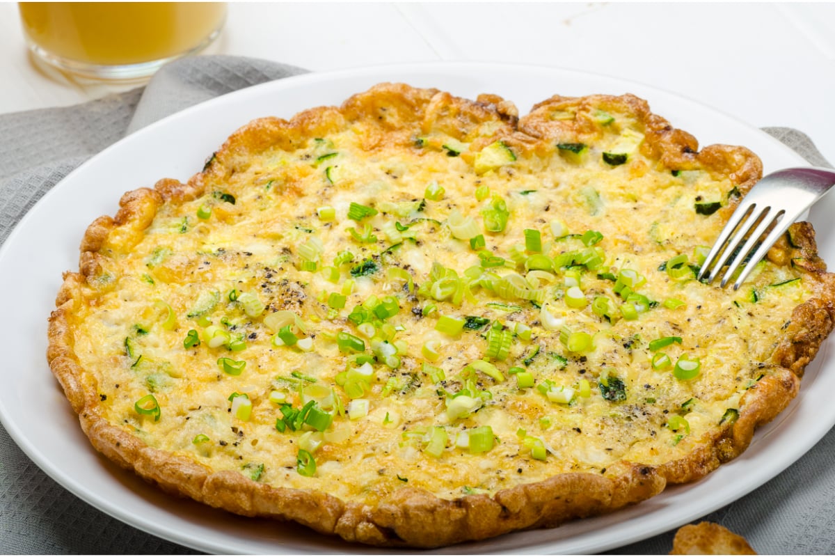 Golden zucchini mozzarella omelette topped with chopped scallions and black pepper, served on a white plate with a fork at the edge.