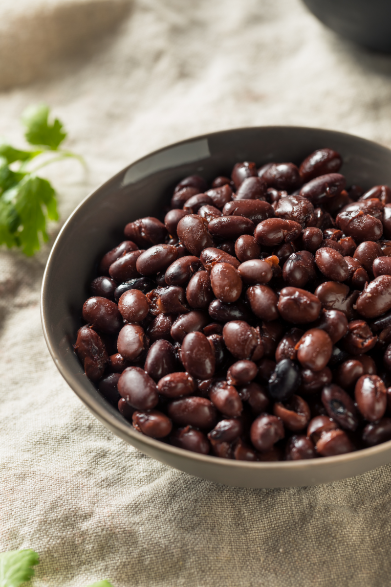 Black beans in a bowl for chicken burrito bowls, shown in a close-up overhead view on a neutral linen cloth with a small sprig of cilantro nearby.
