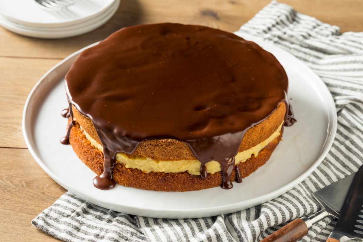 Whole Boston cream pie on a white plate, filled with vanilla pastry cream and topped with glossy chocolate glaze dripping down the sides.