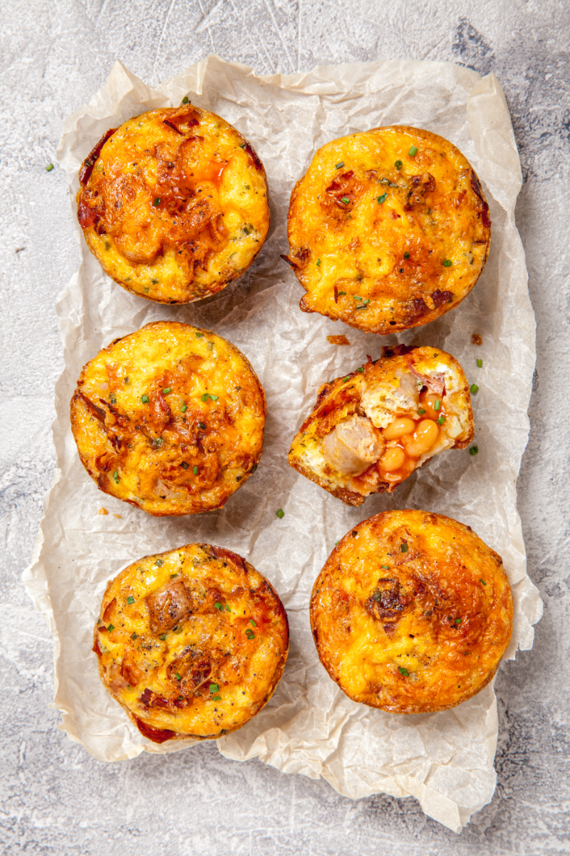Overhead view of golden egg muffins on parchment paper, with one cut open to show a hearty filling and fluffy baked interior.