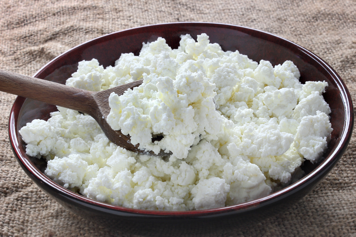 Bowl of creamy cottage cheese with a wooden spoon, shown close up on a rustic fabric surface.