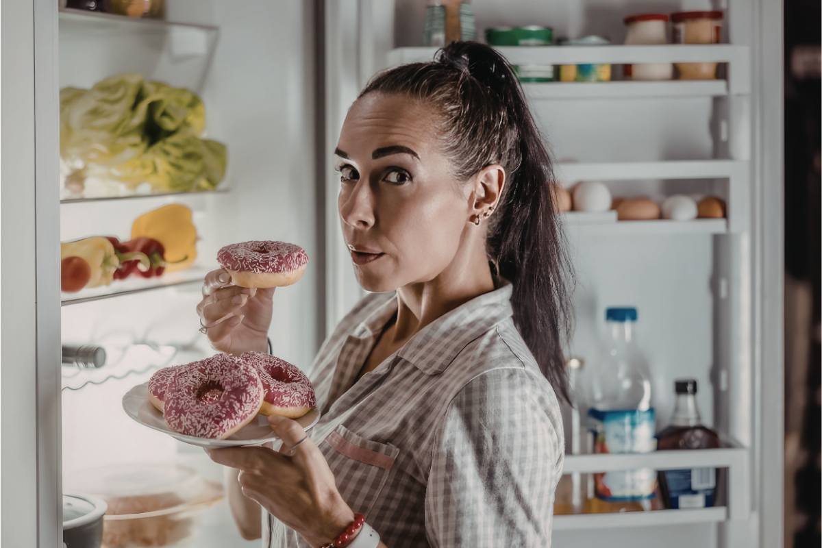 Woman standing at an open refrigerator at night holding pink-frosted donuts, illustrating late-night snacking, sugar before bed, and sleep disruption.