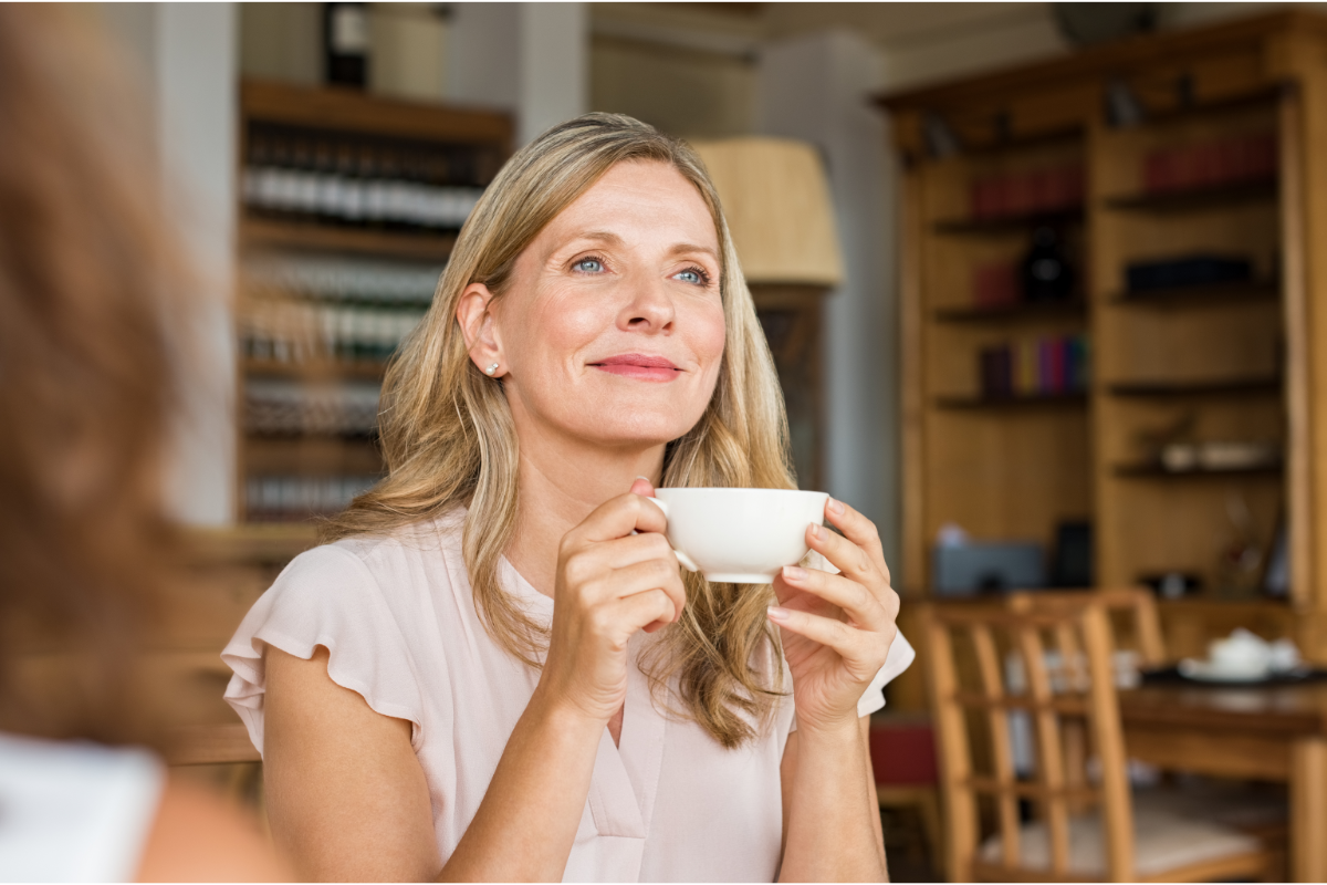 Woman with blonde hair holding a white cup and smiling softly while seated in a warm, cozy café or dining space with wooden shelves in the background.
