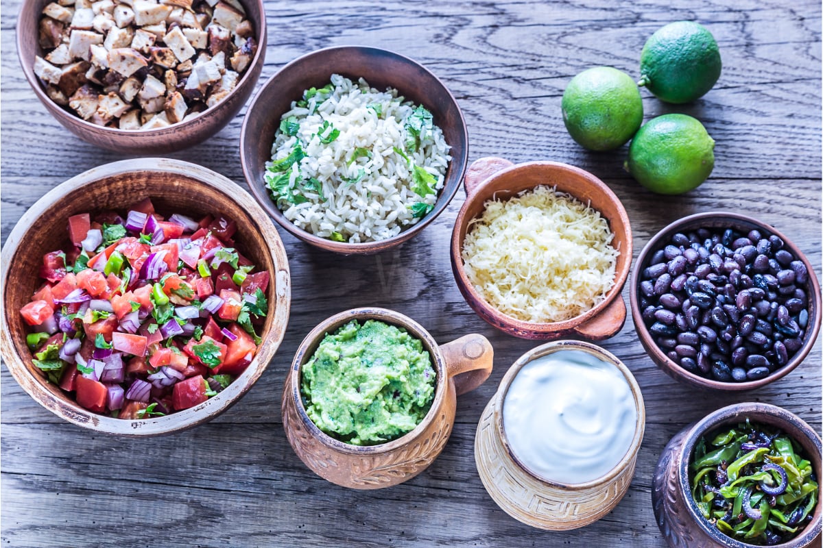 Close-up of burrito bowl ingredients in rustic bowls, including pico de gallo, cilantro lime rice, shredded cheese, black beans, guacamole, sour cream, seasoned chicken, and sautéed peppers with fresh limes.