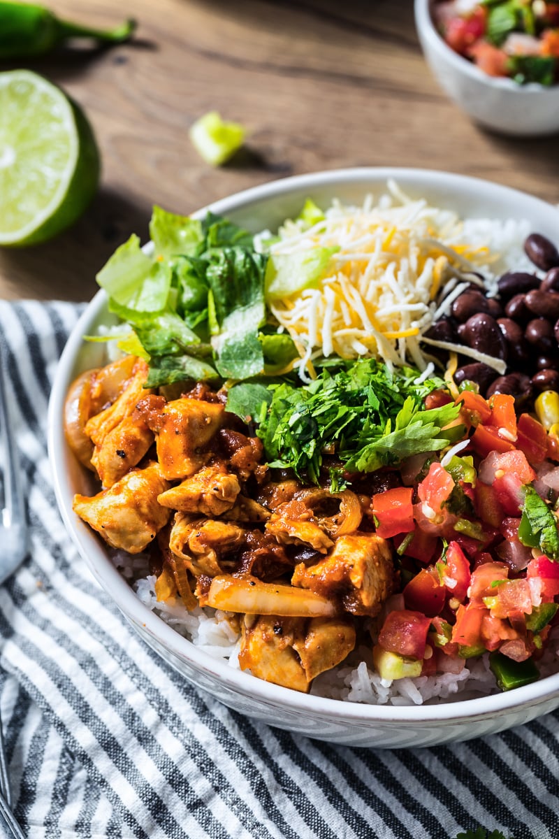 High protein chicken burrito bowl with seasoned chicken, white rice, black beans, shredded cheese, romaine, cilantro, and pico de gallo in a close-up view with lime in the background.