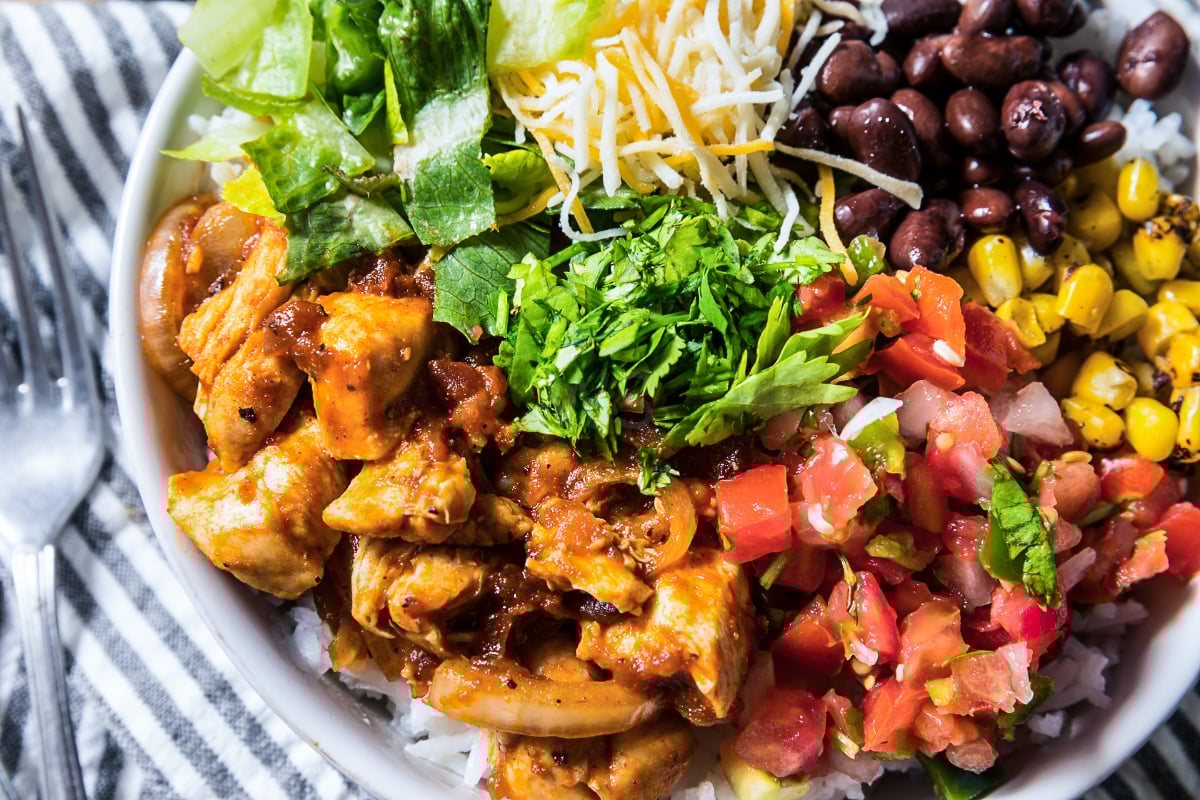 Close-up of a high protein chicken burrito bowl with seasoned chicken, black beans, charred corn, shredded cheese, chopped romaine, cilantro, pico de gallo, and white rice.