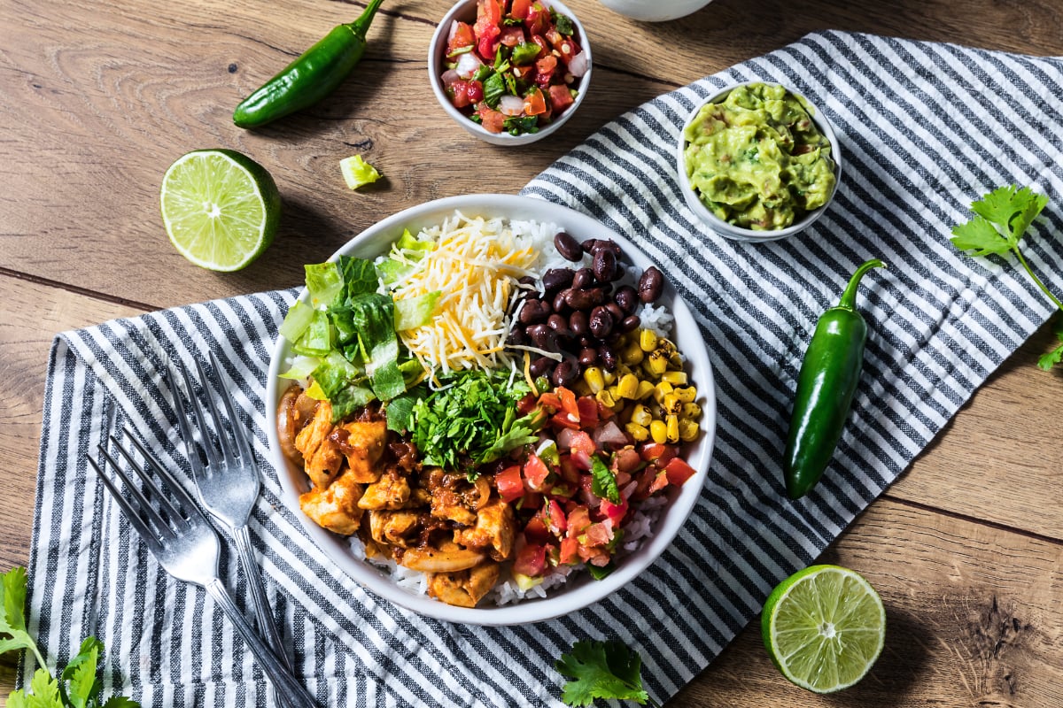 High protein chicken burrito bowl with seasoned chicken, white rice, black beans, corn, shredded cheese, romaine, pico de gallo, guacamole, jalapeños, lime, and cilantro in an overhead view on a rustic wooden table.
