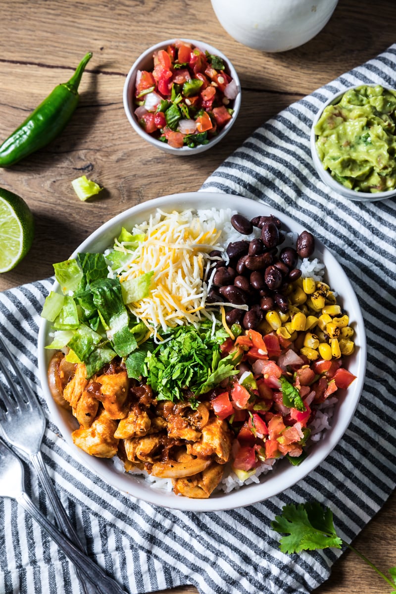 Overhead view of a high protein chicken burrito bowl with seasoned chicken, white rice, black beans, corn, shredded cheese, romaine, pico de gallo, cilantro, guacamole, jalapeño, and lime on a rustic table.
