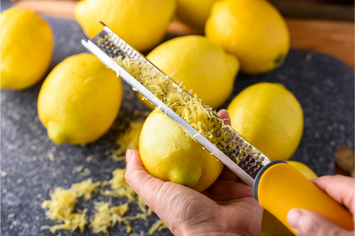 Hand zesting a fresh lemon with a microplane over a dark surface, surrounded by whole lemons and loose lemon zest.