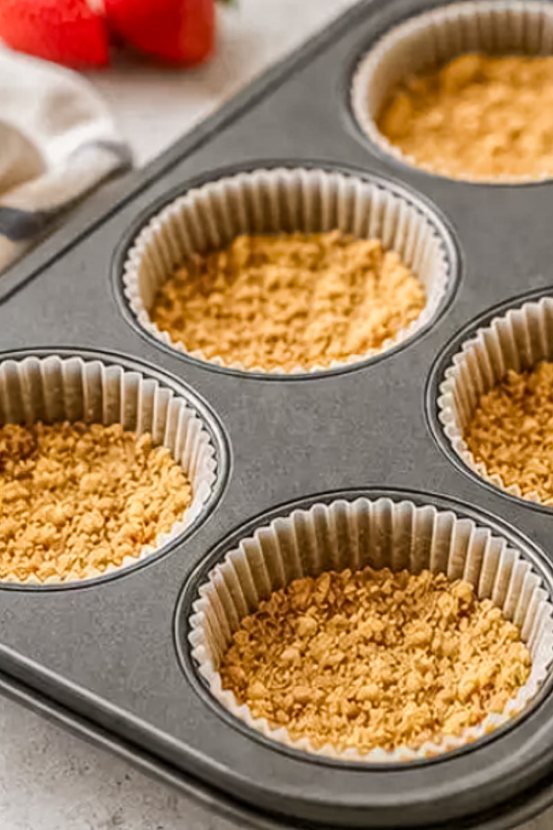 Pressed graham cracker cheesecake crusts inside cupcake liners in a muffin tin.