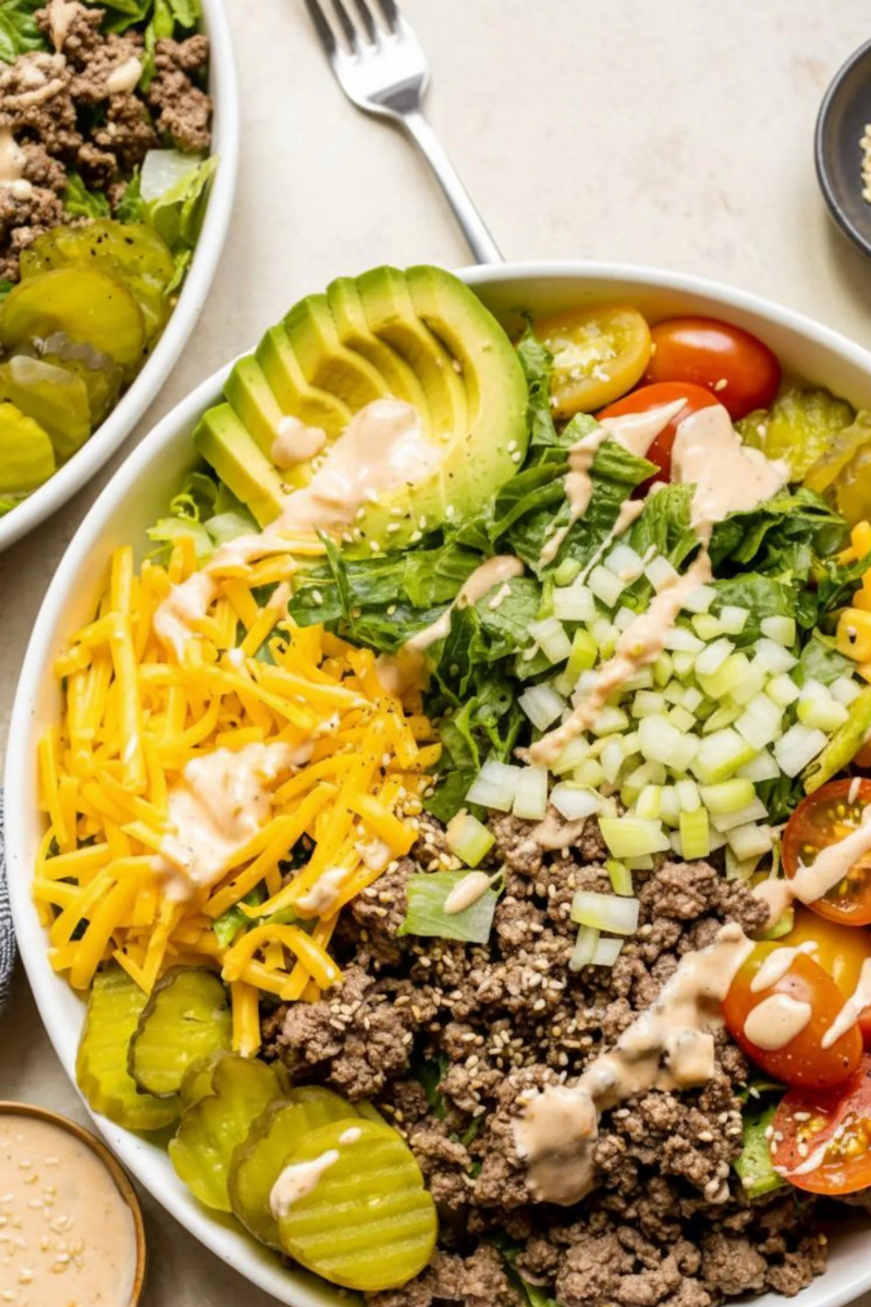 Close-up of a high protein cheeseburger salad bowl with seasoned ground beef, cheddar cheese, avocado, diced onions, tomatoes, pickles, lettuce, spinach, and creamy burger sauce.