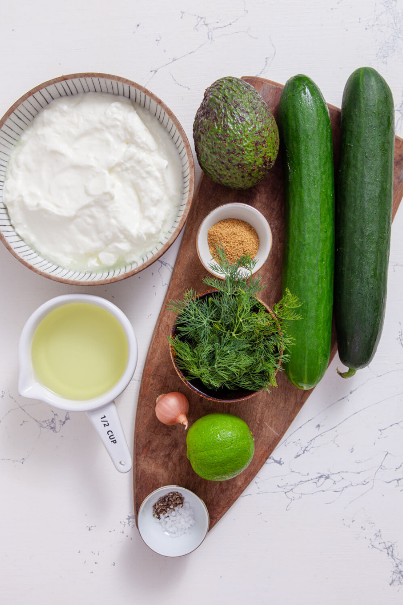 Fresh ingredients for chilled cucumber avocado soup including cucumbers, avocado, Greek yogurt, dill, lime, shallot, vegetable broth, cumin, salt, and pepper arranged on a wooden board.