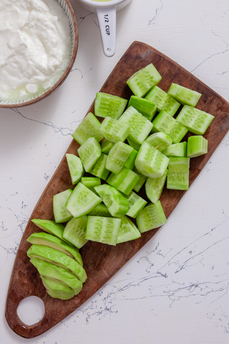 Chopped cucumbers and sliced avocado prepared on a wooden cutting board for chilled cucumber avocado soup.
