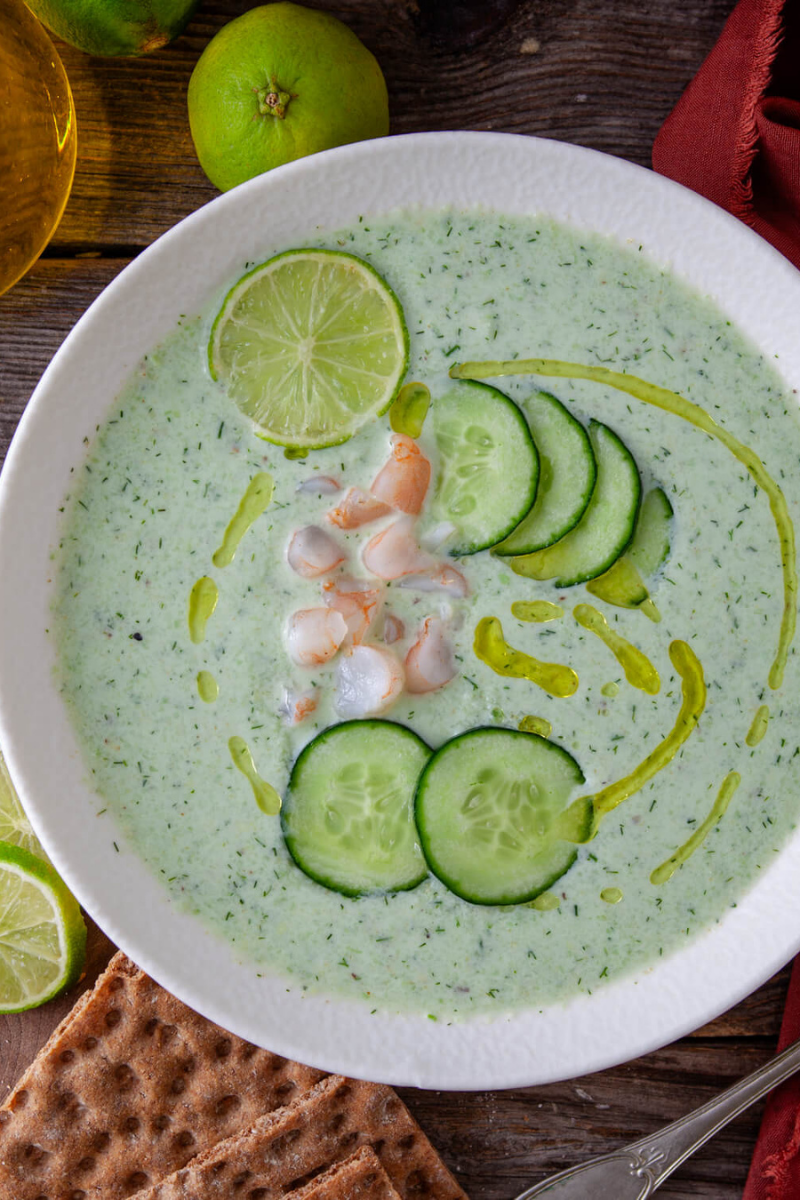 Chilled cucumber avocado soup garnished with cucumber slices, lime, olive oil, dill, and shrimp in a white serving bowl.
