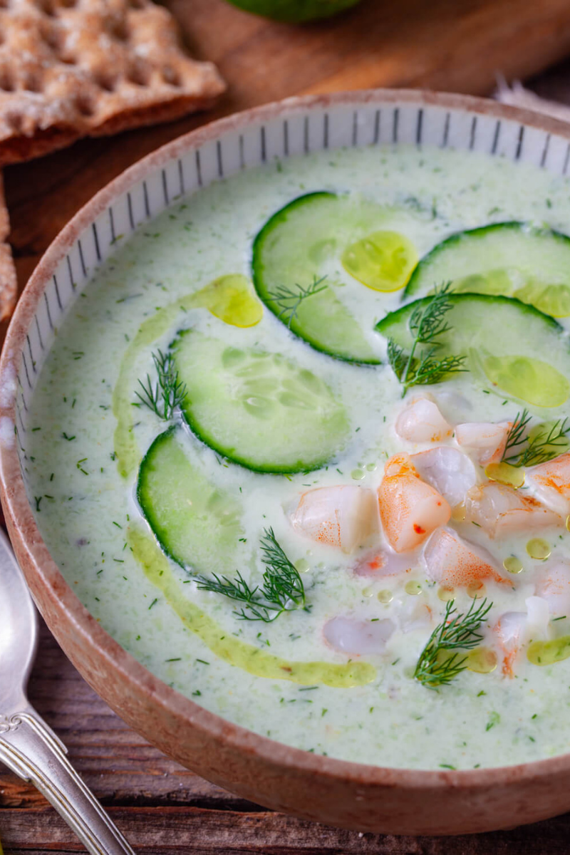 Close-up of chilled cucumber avocado soup with shrimp, cucumber slices, fresh dill, and olive oil drizzle in a rustic serving bowl.