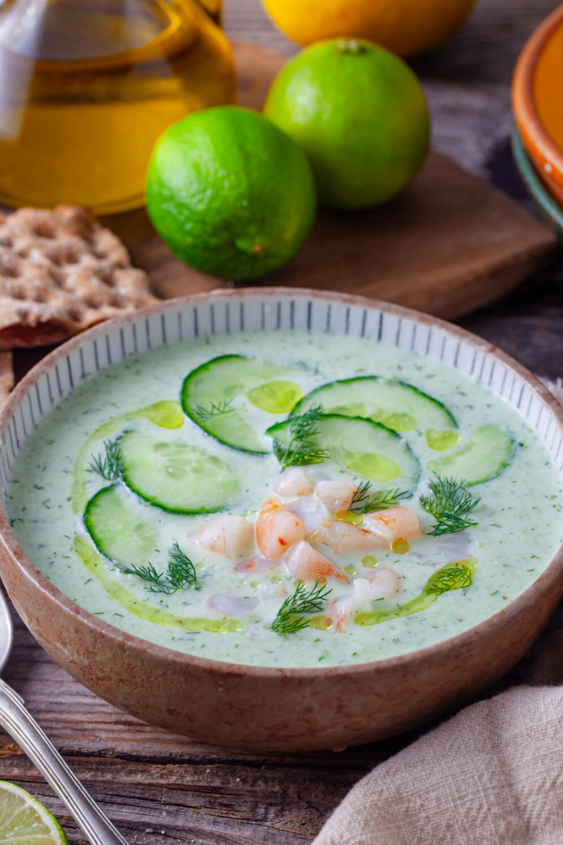 Chilled cucumber avocado soup topped with cucumber slices, fresh dill, olive oil, lime, and shrimp in a rustic bowl on a wooden table.