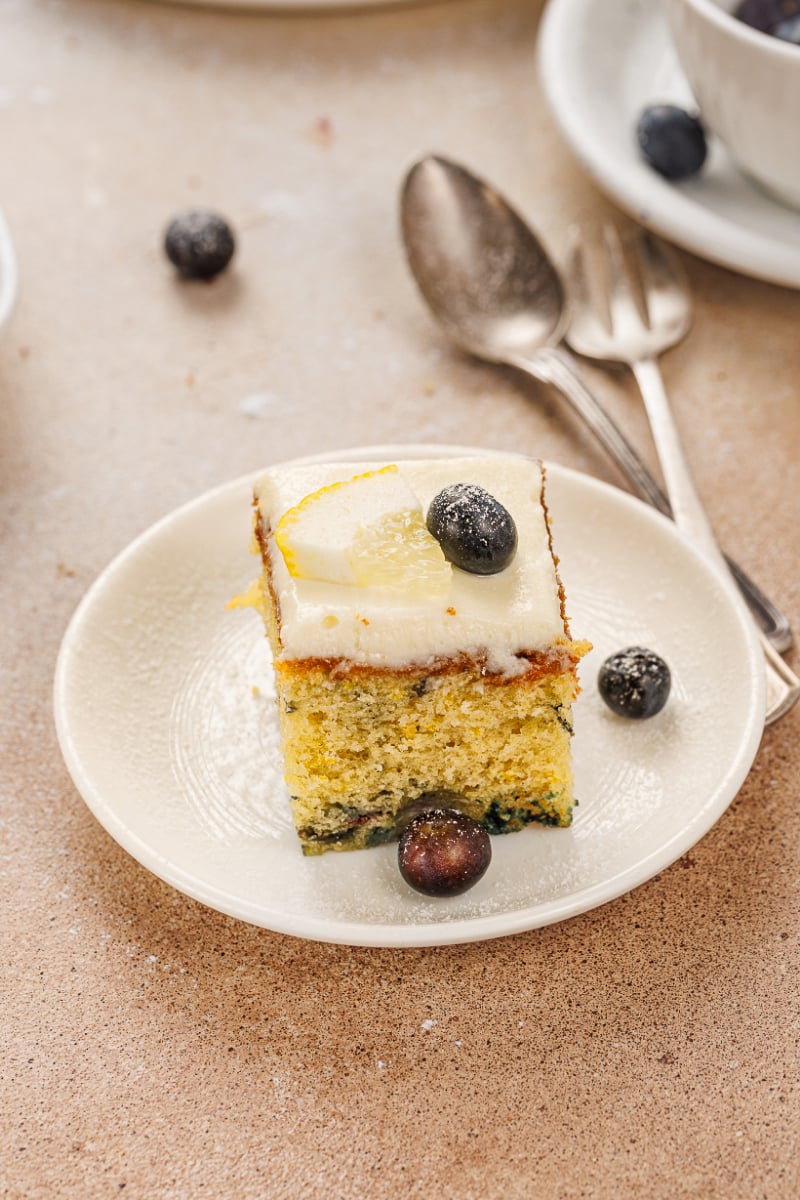 Plated square of lemon blueberry cake with cream cheese frosting, fresh blueberries, and a lemon slice garnish.
