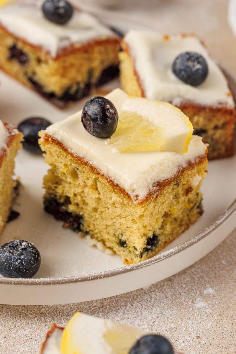 Close-up of lemon blueberry cake squares with cream cheese frosting, fresh blueberries, and a lemon slice garnish on top.