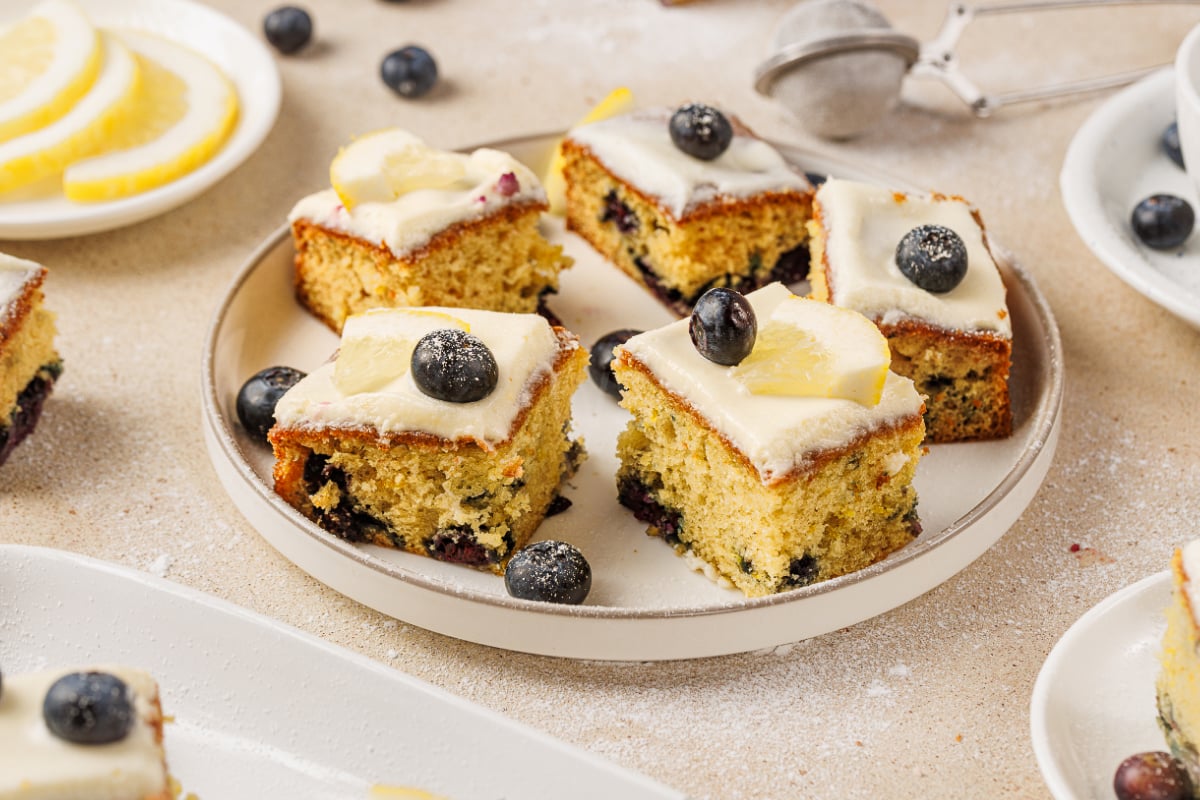 Lemon blueberry cake squares with cream cheese frosting, fresh blueberries, and lemon slices arranged on a plate for serving.