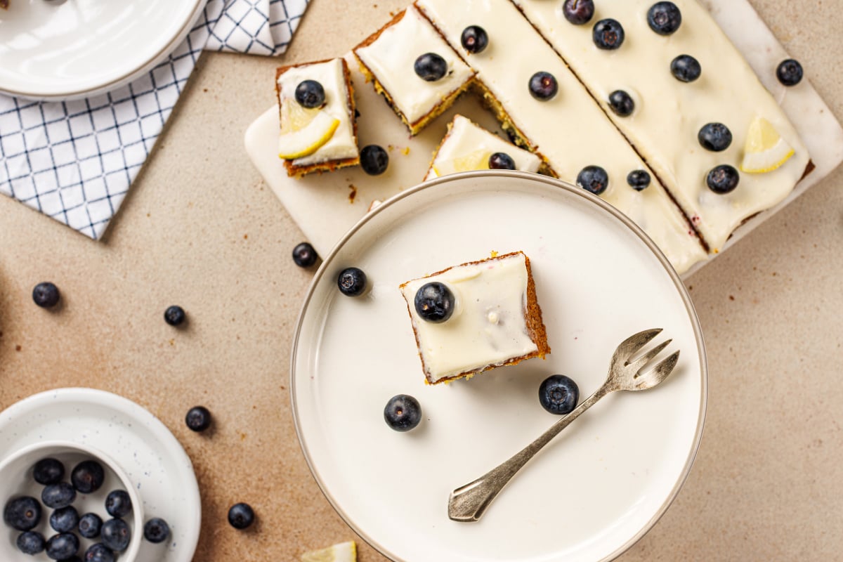 Lemon blueberry cake square served on a plate with cream cheese frosting, fresh blueberries, and the frosted cake sliced in the background.