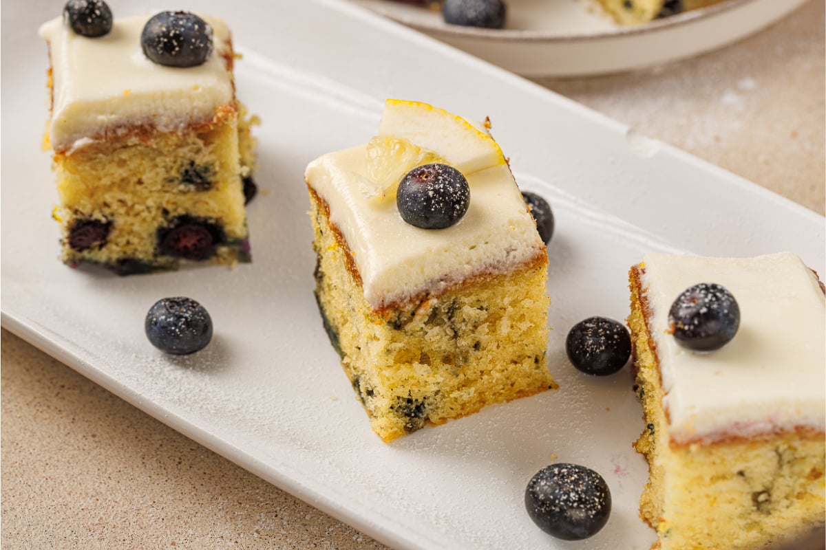 Lemon blueberry cake squares with cream cheese frosting, fresh blueberries, and lemon garnish served on a white platter.