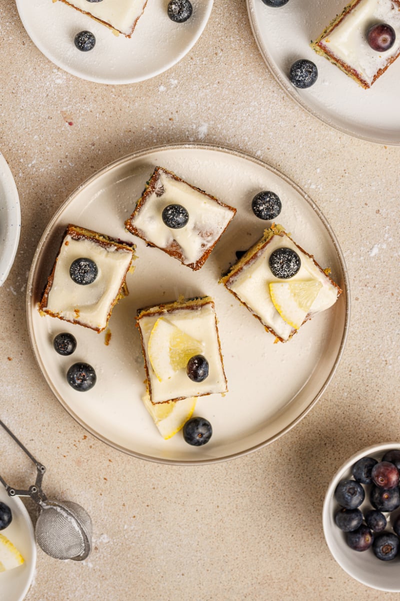 Overhead view of lemon blueberry cake squares with cream cheese frosting, fresh blueberries, and lemon slices arranged on a serving plate.