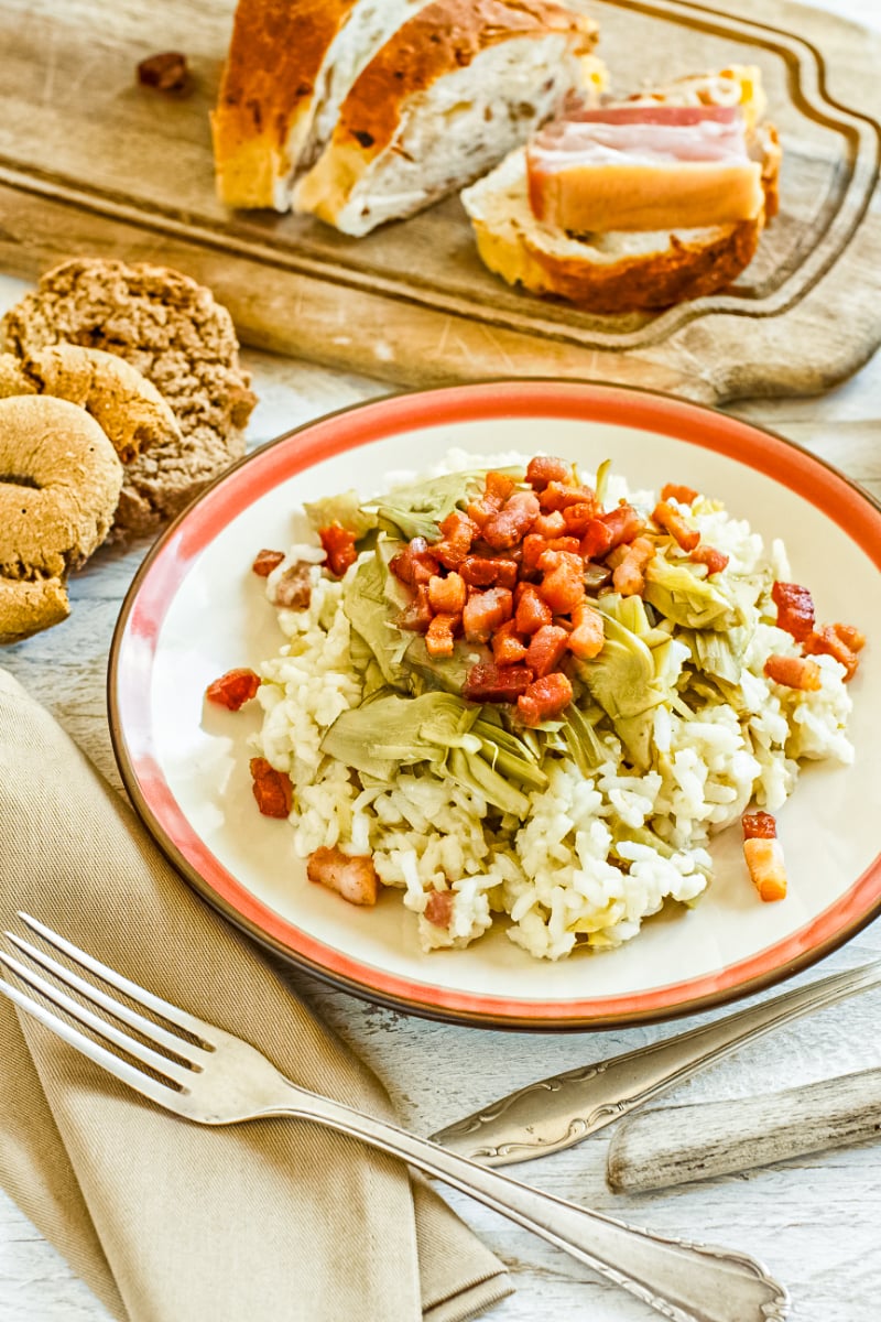Creamy artichoke and bacon risotto served on a white plate with crispy pancetta, sliced bread, and silver forks on a rustic table.