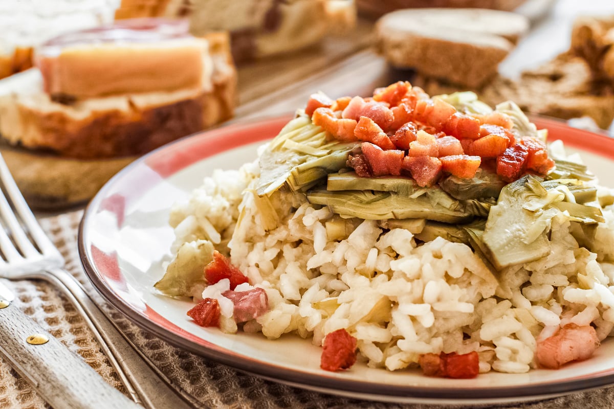 Rustic plate of creamy rice with artichoke hearts, crisp bacon, and bread in the background.
