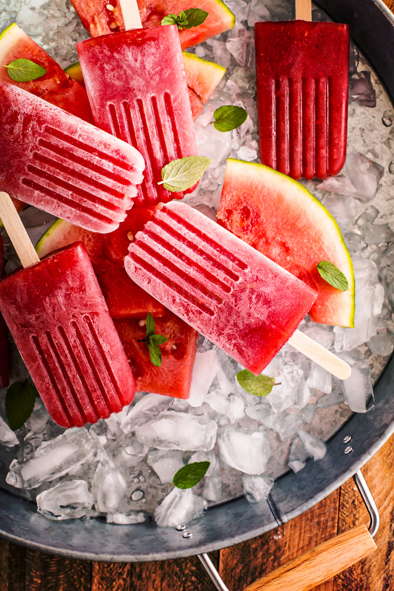 Strawberry watermelon popsicles served over crushed ice with fresh watermelon slices and mint leaves, showing bright red frozen fruit bars for a refreshing homemade summer dessert.