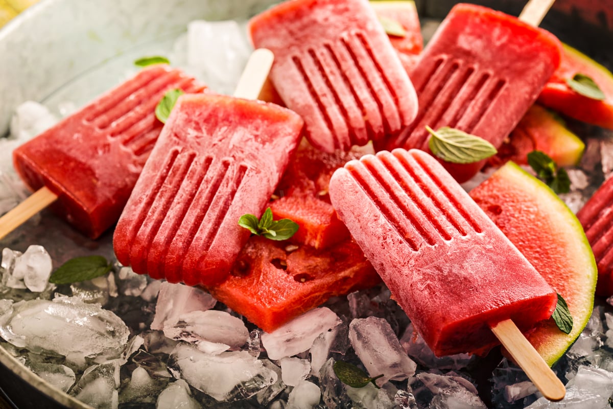 Strawberry watermelon popsicles arranged on crushed ice with fresh watermelon wedges and mint leaves, showcasing vibrant homemade fruit popsicles for a refreshing summer frozen treat.