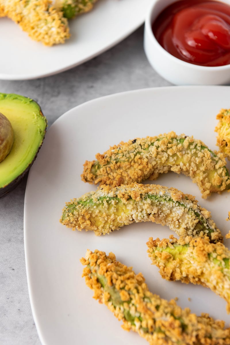 Crispy air fryer avocado fries with golden panko coating, creamy avocado centers, fresh avocado half, and red dipping sauce for an easy vegan appetizer.