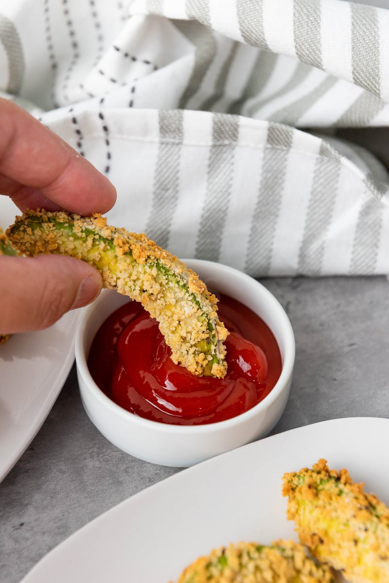 Hand dipping crispy vegan air fryer avocado fries with golden panko coating into red sauce for a crunchy plant-based appetizer or snack.