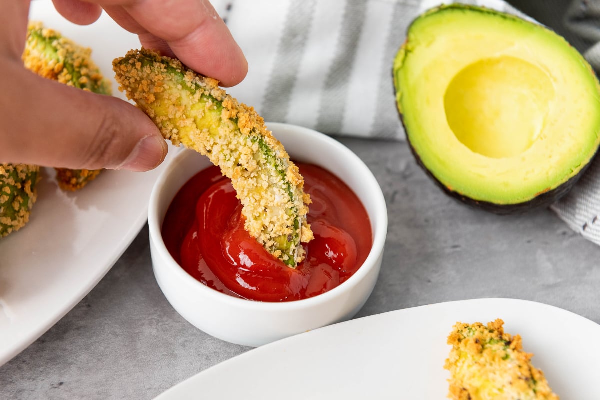 Crispy vegan air fryer avocado fry dipped into red sauce with fresh avocado half in the background for an easy crunchy plant-based appetizer.