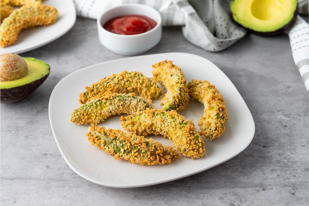 Crispy vegan air fryer avocado fries with golden panko coating, creamy avocado centers, and red dipping sauce served on a white plate for an easy appetizer or snack.