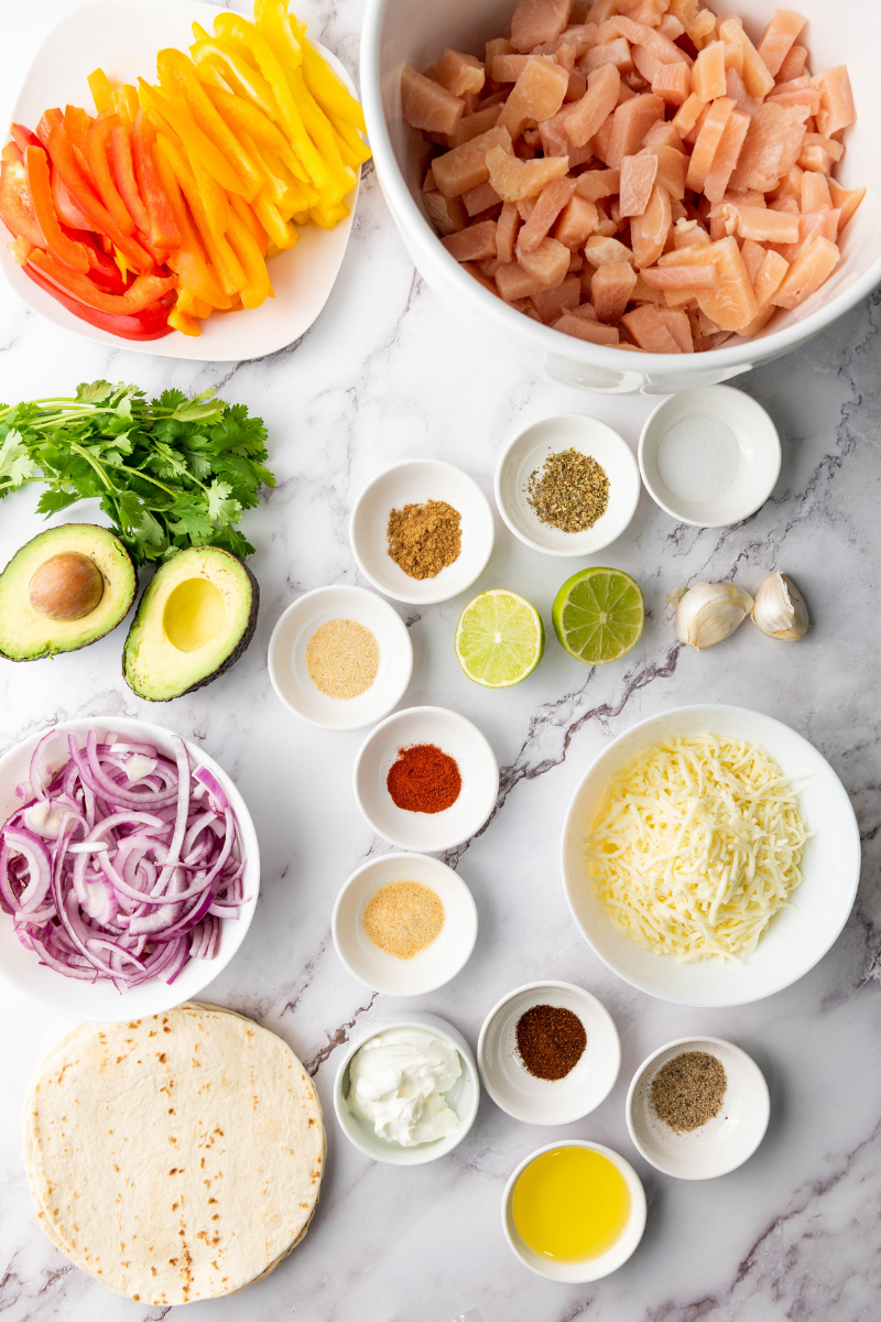 Ingredients for sheet pan chicken fajitas including diced chicken, sliced bell peppers, red onion, spices, garlic, olive oil, tortillas, avocado, lime, and cilantro.