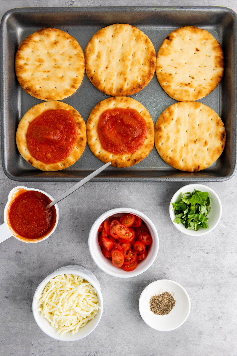 Mini naan rounds spread with pizza sauce on a baking tray with mozzarella cheese, sliced tomatoes, basil, and seasonings ready for assembling Caprese pizzas.