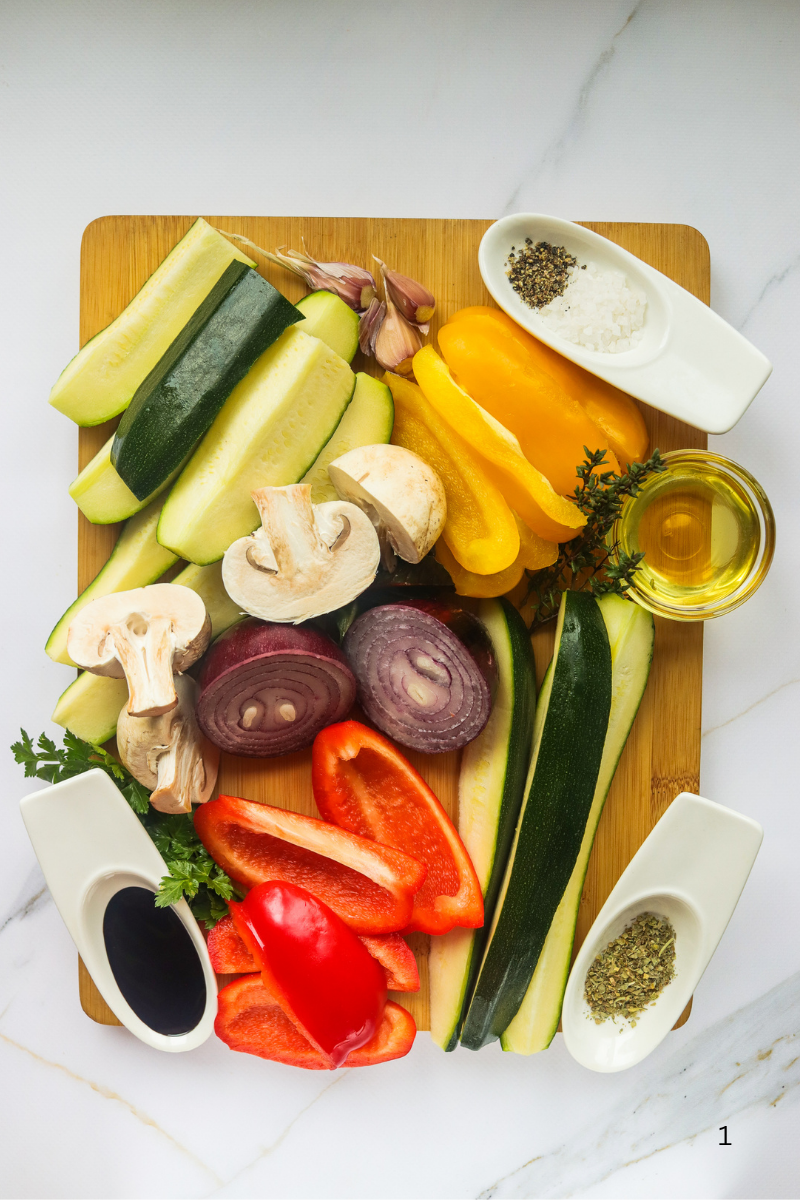 Fresh ingredients for balsamic grilled vegetables including zucchini, bell peppers, red onion, mushrooms, garlic, olive oil, balsamic vinegar, herbs, salt, and pepper on a cutting board.