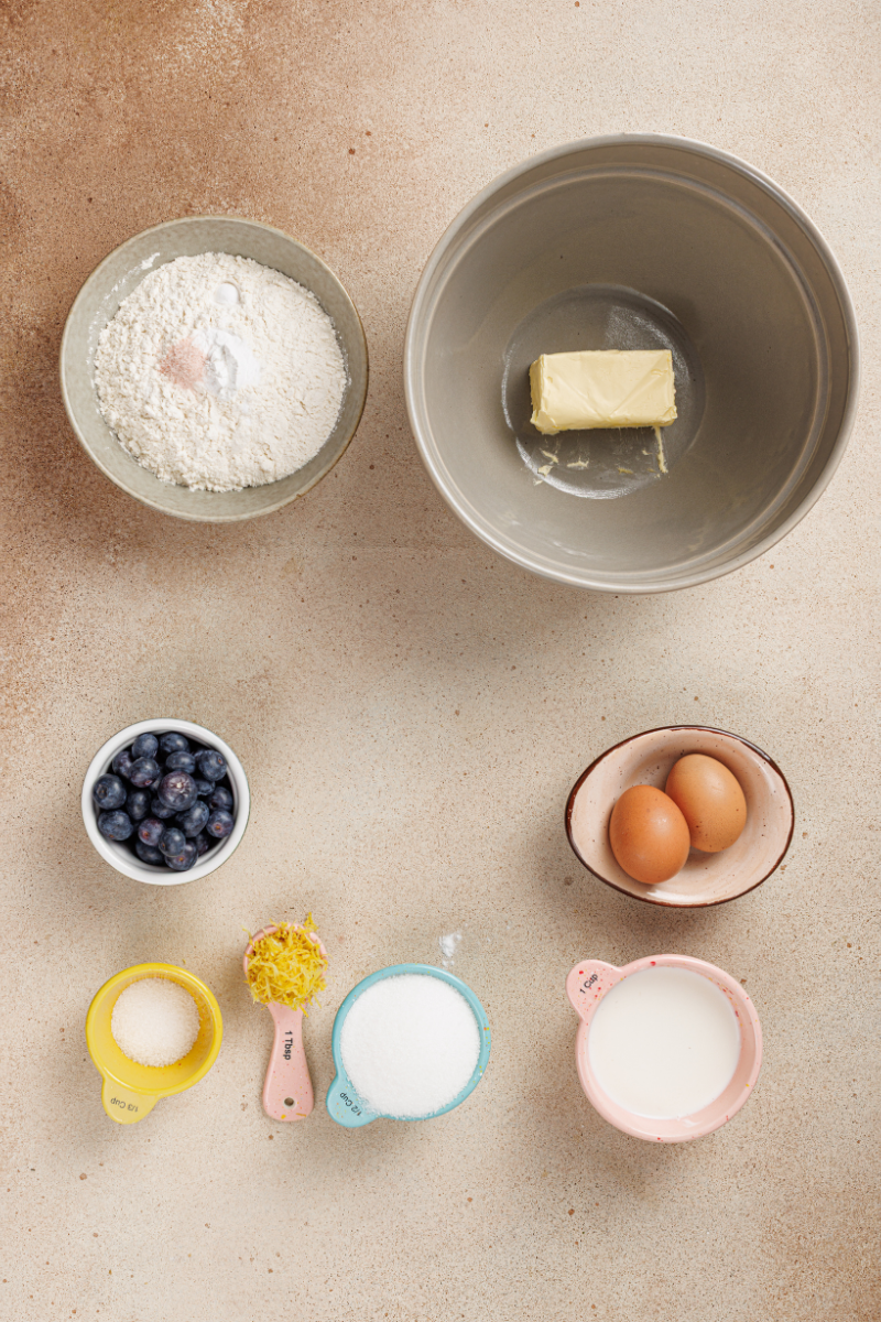 Ingredients for lemon blueberry cake arranged on a counter, including butter, eggs, flour, sugar, buttermilk or yogurt, lemon zest, and fresh blueberries.