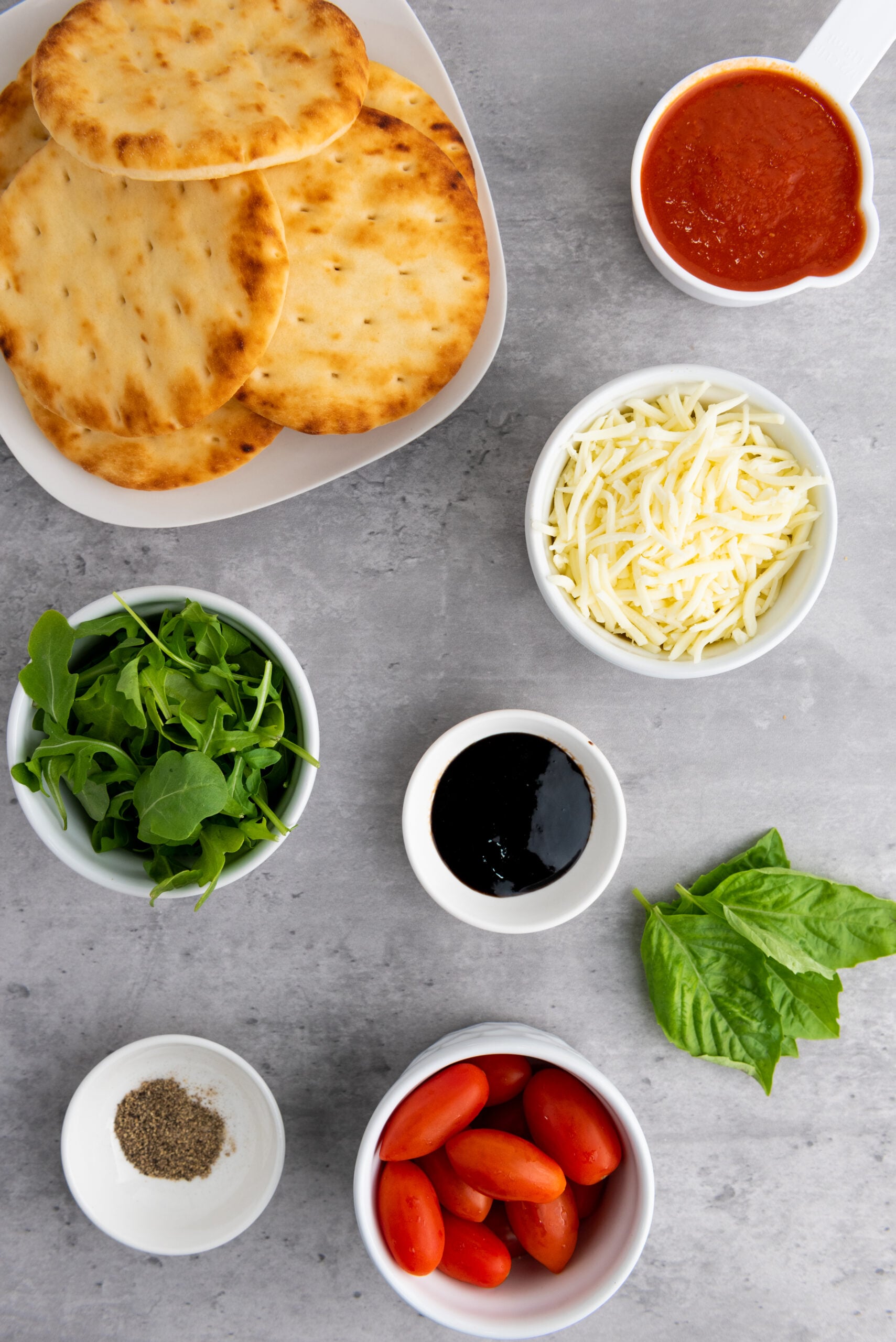Ingredients for mini Caprese naan pizzas including naan bread, mozzarella cheese, pizza sauce, cherry tomatoes, basil, arugula, balsamic glaze, and black pepper arranged on a countertop.
