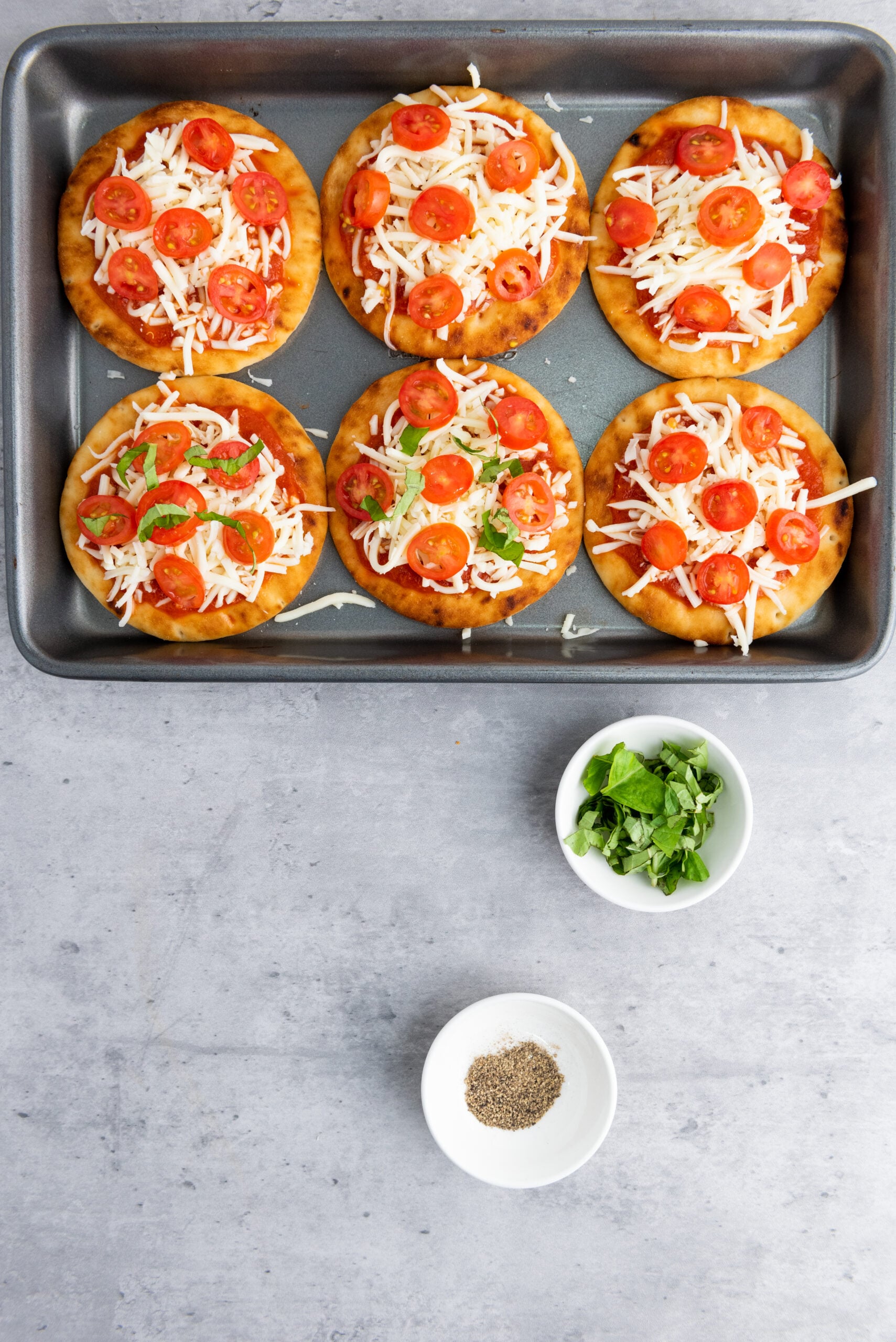 Mini Caprese naan pizzas topped with mozzarella cheese, sliced cherry tomatoes, and fresh basil on a baking tray before oven baking.