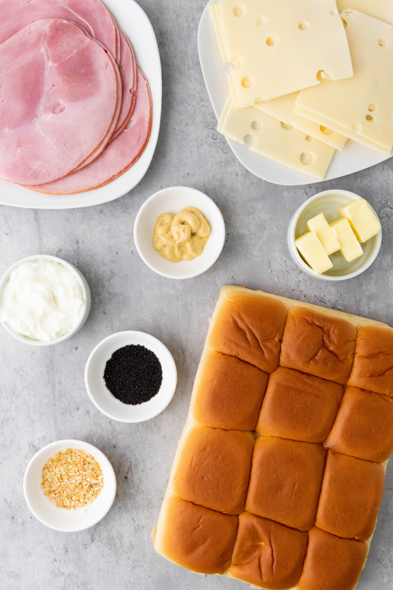 Ingredients for ham and cheese sliders including slider buns, sliced ham, Swiss cheese, mayonnaise, Dijon mustard, butter, minced onion, and poppy seeds.