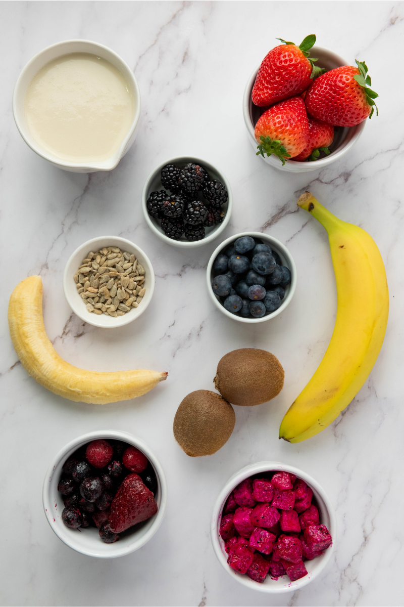 Smoothie bowl ingredients arranged on marble, including banana, dragon fruit, mixed berries, kiwi, strawberries, blueberries, blackberries, sunflower seeds, and milk.