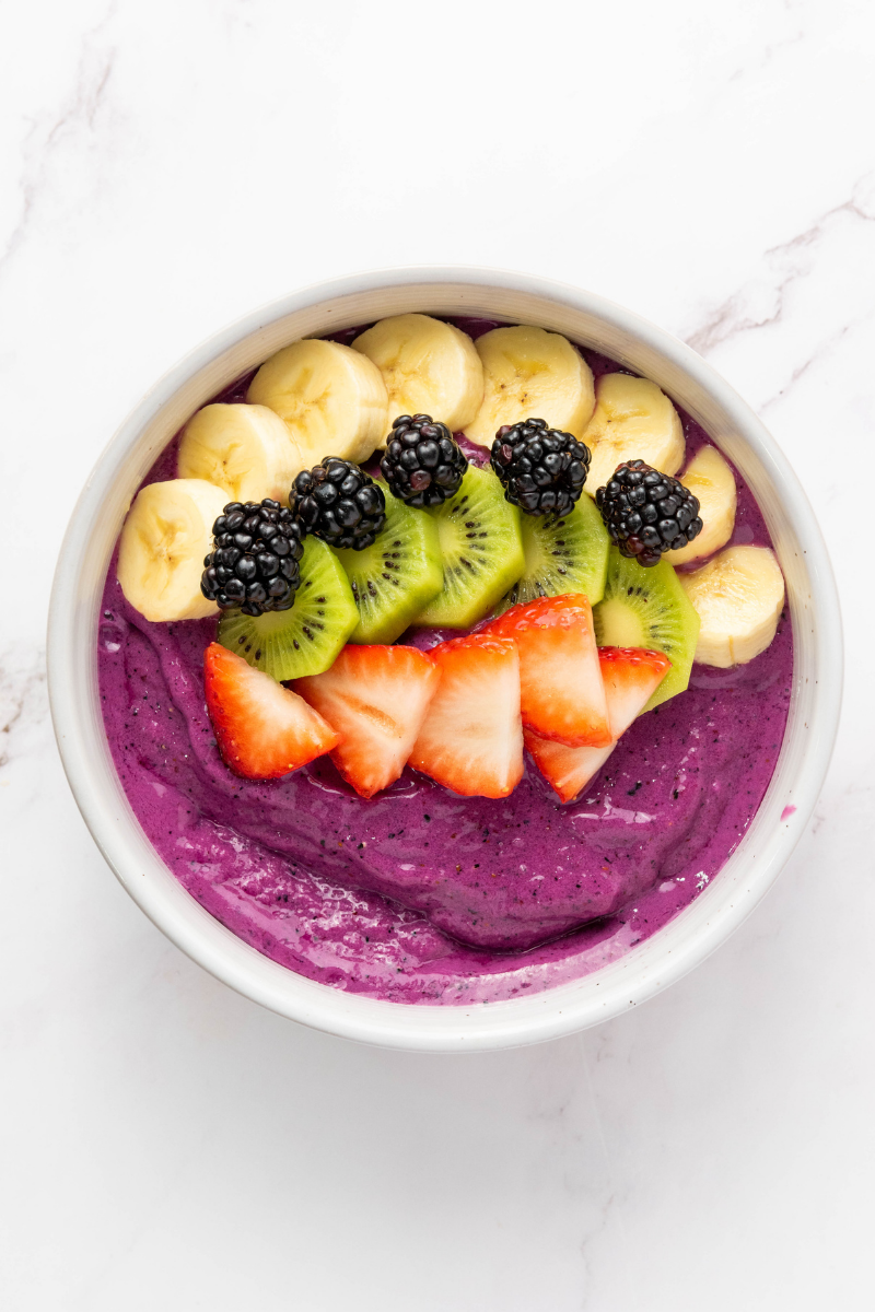 Purple smoothie bowl topped with sliced banana, kiwi, strawberries, and blackberries arranged in colorful rows.
