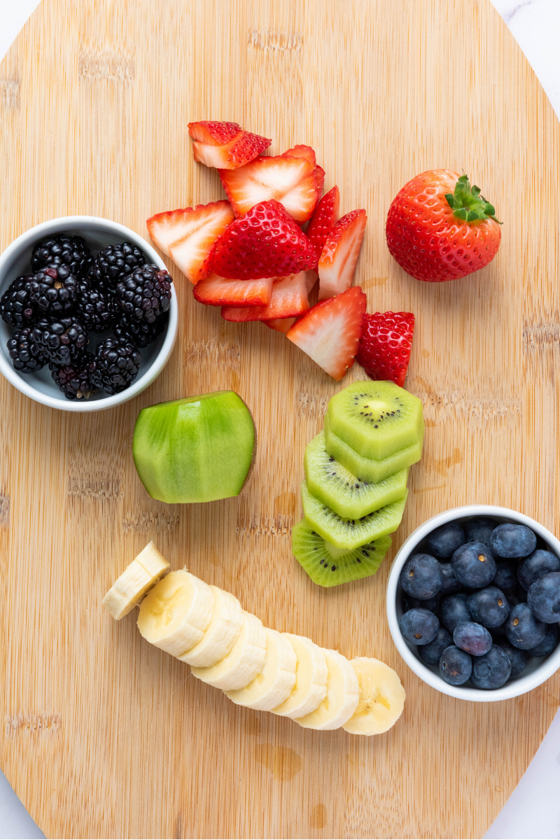 Fresh smoothie bowl toppings prepared on a cutting board, including sliced banana, kiwi, strawberries, blueberries, and blackberries.