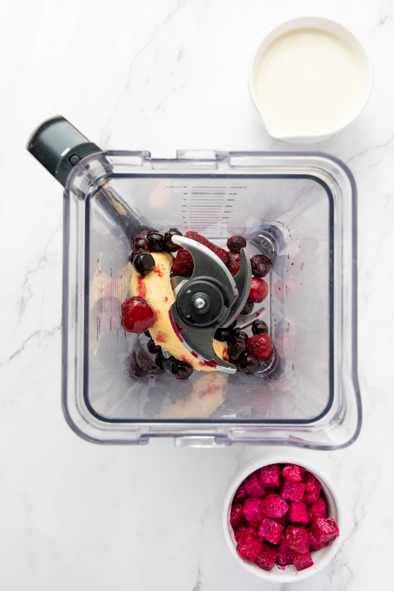 Frozen berries and banana added to a blender for a thick smoothie bowl, with dragon fruit and milk nearby.
