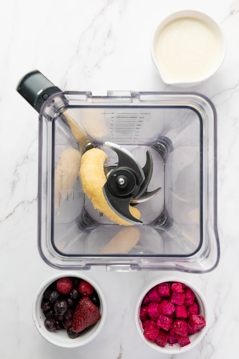 Banana added to a blender as the first step for a smoothie bowl, with frozen berries, dragon fruit, and milk nearby.
