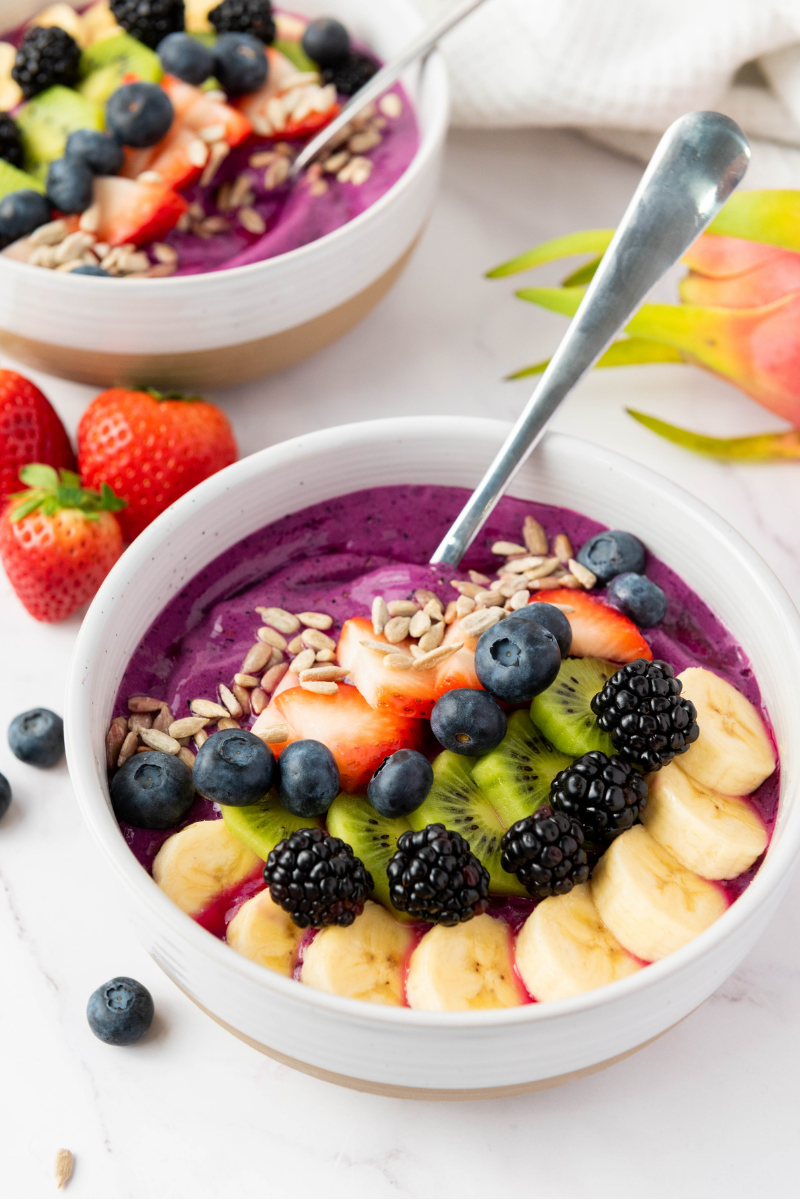 Purple smoothie bowl topped with fresh fruit and sunflower seeds, served with a spoon and a second bowl in the background.