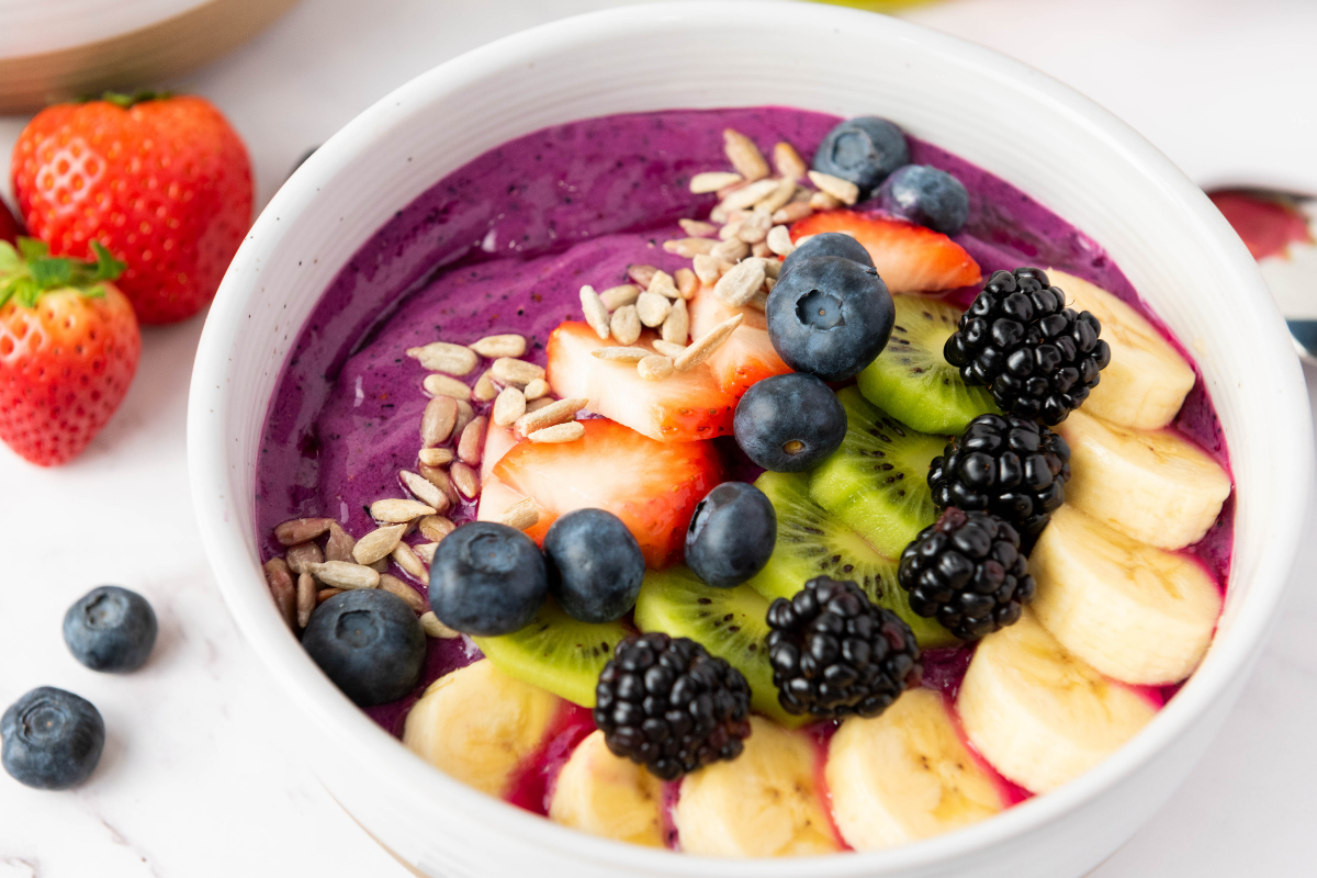 Purple smoothie bowl topped with banana, kiwi, strawberries, blueberries, blackberries, and sunflower seeds, with fresh berries on the side.